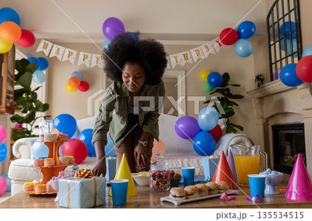 African American woman leaning over table at home organizing cupcakes by gift box and balloons African American woman leaning over table at home organizing cupcakes by gift box and balloons 135534515