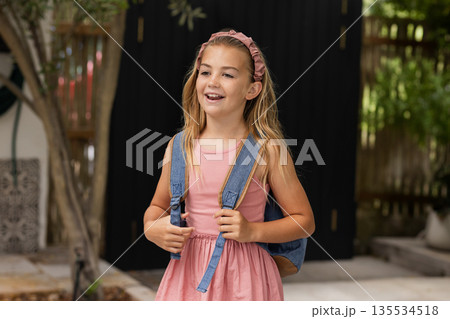 Girl child standing on home patio, holding denim backpack straps, wearing pink headband, smiling Girl child standing on home patio, holding denim backpack straps, wearing pink headband, smiling 135534518