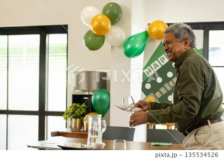 Holding wine glasses middle aged man arranging tableware in dining room, with balloons and banner 135534526