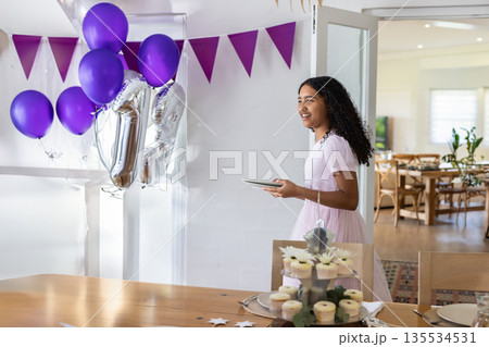 Child girl standing beside table at home holding tablet amid cupcakes and balloons, copy space 135534531