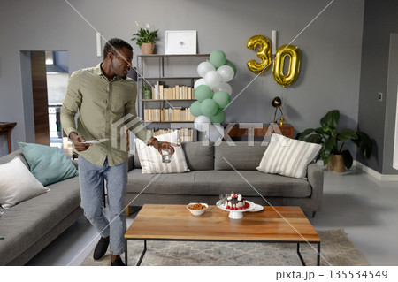 African American man arranging tablet on table in living room with birthday cake and balloons 135534549