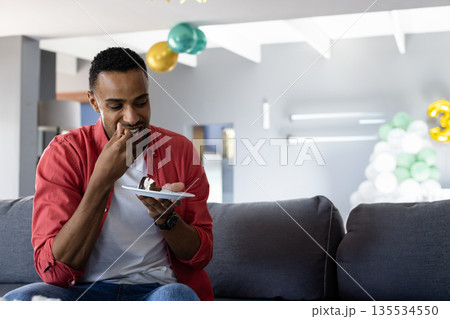 Mid adult African American man sitting on sofa eating cake under balloons at home, copy space 135534550