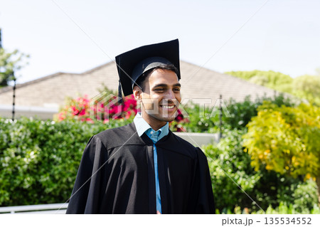 Indian man smiling while standing in front yard wearing black mortarboard cap and gown 135534552