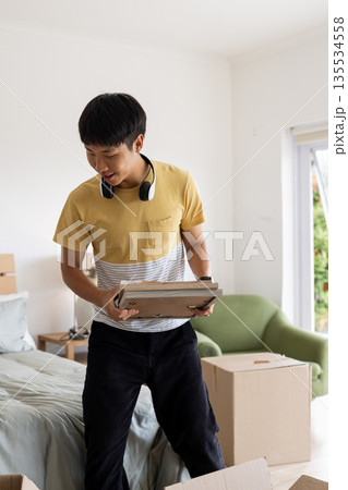 Asian man holding wooden frame while unpacking boxes in bedroom near green armchair with headphones Asian man holding wooden frame while unpacking boxes in bedroom near green armchair with headphones 135534558