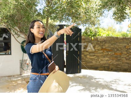 Mid adult Asian woman pointing in sunlit courtyard holding straw hat journal bag and wristwatch 135534568
