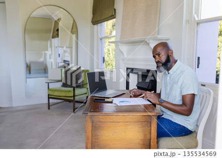 African American man reviewing printed chart in home office at desk with laptop, copy space African American man reviewing printed chart in home office at desk with laptop, copy space 135534569
