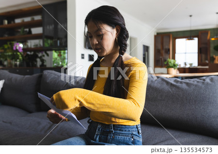 Asian woman holding white envelope sitting on grey sofa with potted plants in modern living area 135534573