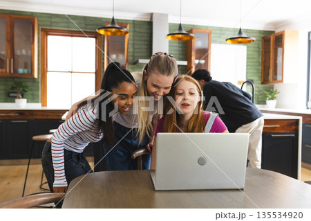 Teen friends gathering around laptop in kitchen, sharing excitement and laughter 135534920
