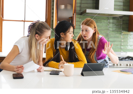 Teen girls collaborating on school project in kitchen, using tablet and notes Teen girls collaborating on school project in kitchen, using tablet and notes 135534942