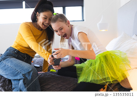 Teen girls laughing and sharing exciting moment on smartphone in bedroom Teen girls laughing and sharing exciting moment on smartphone in bedroom 135535011