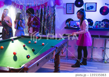 Teen playing pool at birthday party, friends enjoying music and snacks nearby Teen playing pool at birthday party, friends enjoying music and snacks nearby 135535063