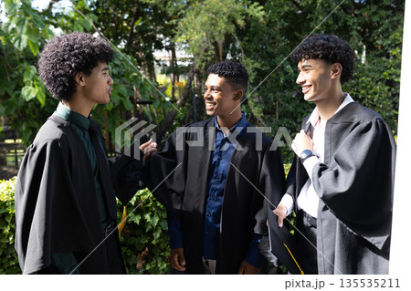 Teen graduates in gowns celebrating outdoors, sharing joyful conversation and smiles Teen graduates in gowns celebrating outdoors, sharing joyful conversation and smiles 135535211