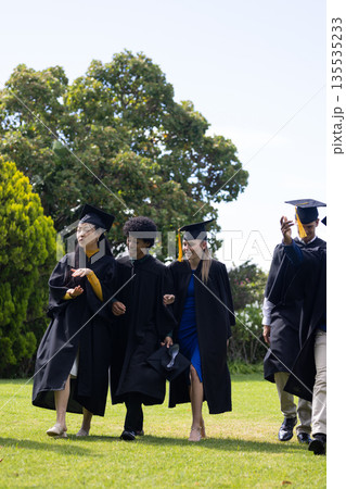 Graduating students in caps and gowns walking joyfully on grassy field 135535233