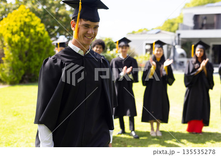 Graduate in cap and gown walking confidently on campus, friends clapping behind 135535278