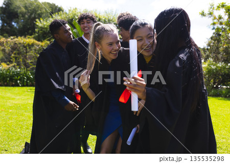 Teen graduates in gowns celebrating outdoors, holding diplomas and smiling joyfully 135535298