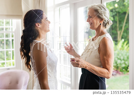 Bride and older woman sharing joyful moment near window on wedding day Bride and older woman sharing joyful moment near window on wedding day 135535479