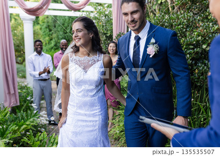 Bride and groom walking joyfully down garden aisle, guests applauding happily 135535507