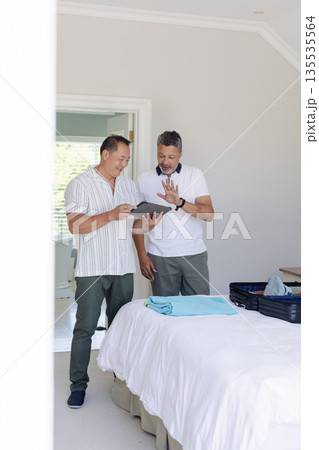 Two men in bedroom packing suitcase and discussing travel plans using tablet Two men in bedroom packing suitcase and discussing travel plans using tablet 135535564