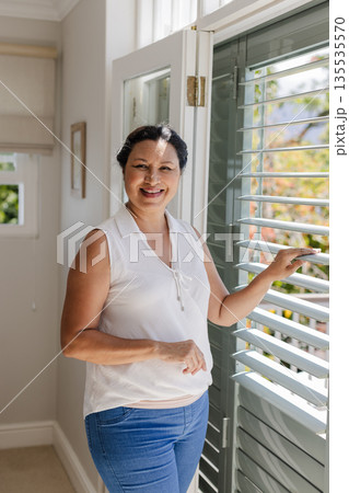 Senior woman smiling while opening window blinds at home, enjoying sunlight Senior woman smiling while opening window blinds at home, enjoying sunlight 135535570