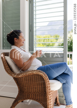 Relaxing at home, woman enjoying coffee while sitting in wicker chair by window Relaxing at home, woman enjoying coffee while sitting in wicker chair by window 135535588