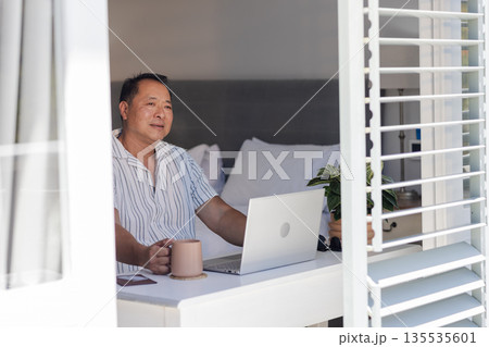 Asian man enjoying coffee while using laptop at home near window Asian man enjoying coffee while using laptop at home near window 135535601