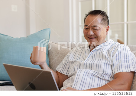 Senior man enjoying coffee while using laptop at home, smiling contentedly Senior man enjoying coffee while using laptop at home, smiling contentedly 135535612