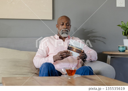 Senior man reading book on sofa at home, enjoying relaxing tea time 135535619