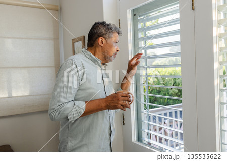 Senior man holding coffee, looking out window at home, appearing thoughtful 135535622