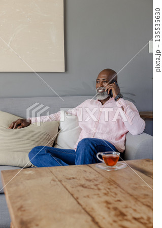 Senior man in pink shirt relaxing on sofa, talking on smartphone with tea nearby 135535630