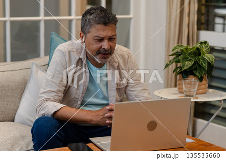 Senior man using laptop at home, wearing earbuds, focused on video call 135535660
