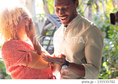 African American man surprising friend with gift outdoors, both looking delighted African American man surprising friend with gift outdoors, both looking delighted 135535782
