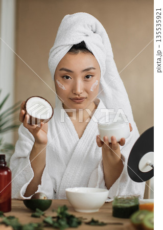 A young Asian woman in a white bathrobe and towel applies a natural face cream with coconut extract for skincare A young Asian woman in a white bathrobe and towel applies a natural face cream with coconut extract for skincare 135535921