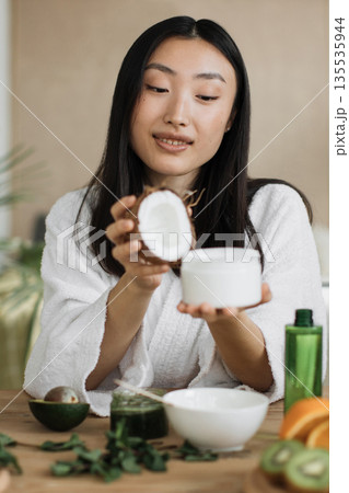 A young woman in a white robe holds a halved coconut and a jar of cream, surrounded by natural ingredients like avocado and mint, suggesting a homemade beauty treatment A young woman in a white robe holds a halved coconut and a jar of cream, surrounded by natural ingredients like avocado and mint, suggesting a homemade beauty treatment 135535944