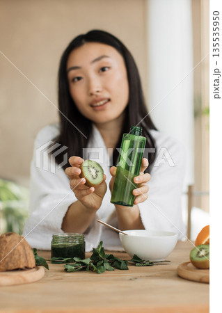 A young Asian woman in a white robe holds a kiwi slice and a green bottle of serum, surrounded by ingredients for homemade beauty products A young Asian woman in a white robe holds a kiwi slice and a green bottle of serum, surrounded by ingredients for homemade beauty products 135535950
