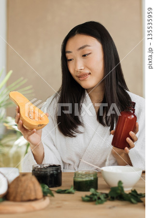A young woman in a white robe holds a halved butternut squash and a bottle of serum, surrounded by natural ingredients for homemade skincare 135535959