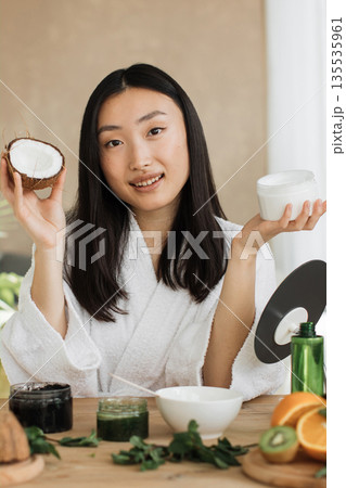 A young Asian woman in a white robe holds a halved coconut and a jar of cream, surrounded by ingredients for homemade beauty products 135535961