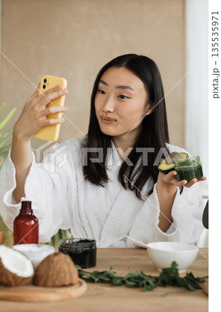 A young Asian woman in a white bathrobe films her natural skincare routine with avocado and coconut ingredients on her phone A young Asian woman in a white bathrobe films her natural skincare routine with avocado and coconut ingredients on her phone 135535971