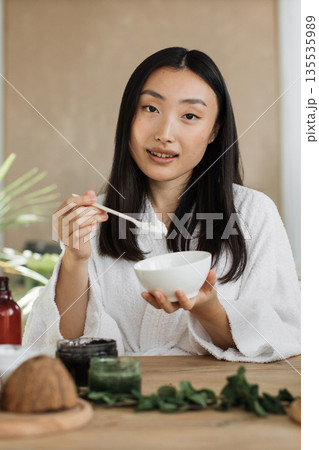 A young Asian woman in a white bathrobe holds a bowl and spoon, preparing to apply a natural skincare product A young Asian woman in a white bathrobe holds a bowl and spoon, preparing to apply a natural skincare product 135535989