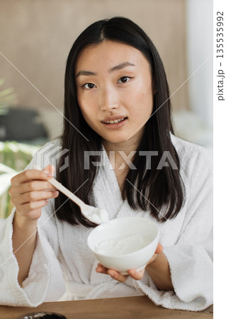 A young Asian woman in a white bathrobe holds a bowl and spoon, preparing to apply a skincare product to her face 135535992