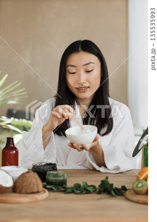 A young Asian woman in a white bathrobe mixes ingredients for a homemade facial mask, surrounded by natural elements like coconut and fruit A young Asian woman in a white bathrobe mixes ingredients for a homemade facial mask, surrounded by natural elements like coconut and fruit 135535993