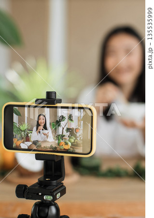 A young woman films herself preparing a healthy meal for her online audience, surrounded by fresh ingredients and plants A young woman films herself preparing a healthy meal for her online audience, surrounded by fresh ingredients and plants 135535999