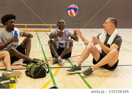 Diverse male partners sitting on sports hall floor tossing red white blue volleyball with towel 135536104