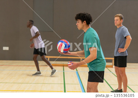 Diverse male volleyball players holding red-white-blue ball, moving into position on gym court 135536105