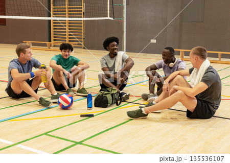 Diverse male teammates sitting on court chatting, hydrating with water bottles beside volleyball 135536107
