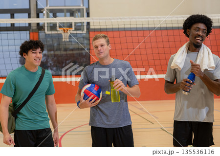 Diverse male friends standing on gym wood floor court holding volleyball water bottles, gym bag 135536116