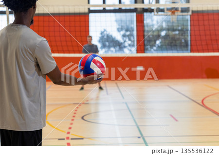 Diverse male volleyball teammates training in gym, holding volleyball next to net and hoop Diverse male volleyball teammates training in gym, holding volleyball next to net and hoop 135536122