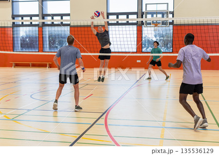 Diverse male partners in sportswear spiking volleyball over net at gym with backboard, court lines 135536129