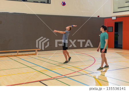 Two male volleyball players serving ball, watching in gym on court with bench, boundary lines 135536141