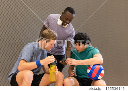 Diverse male partners checking smartphone on bench at gym with water bottle volleyball headphones 135536143