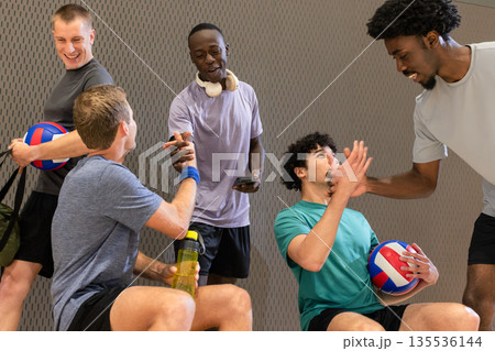 Diverse male volleyball partners resting in sports hall holding volleyballs and water bottle 135536144